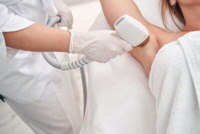 Top view cropped head of female lying on couch while professional is using device for removing hair on armpit